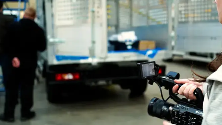 A camera filming two workers next to a van.