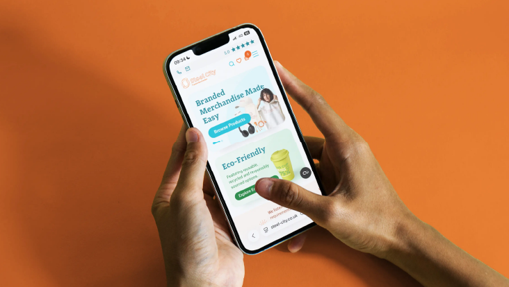 A pair of hands holding a phone displaying the Steel City Website against an orange background.